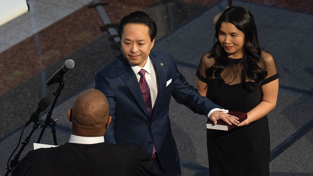 Swearing in ceremony showing the back of the judge's head and the new DA facing the judge, smiling, with his hand on a Bible, and the DA's wife standing beside him.