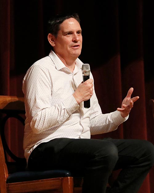 A man in a white shirt sits on stage, holding a microphone and talking.