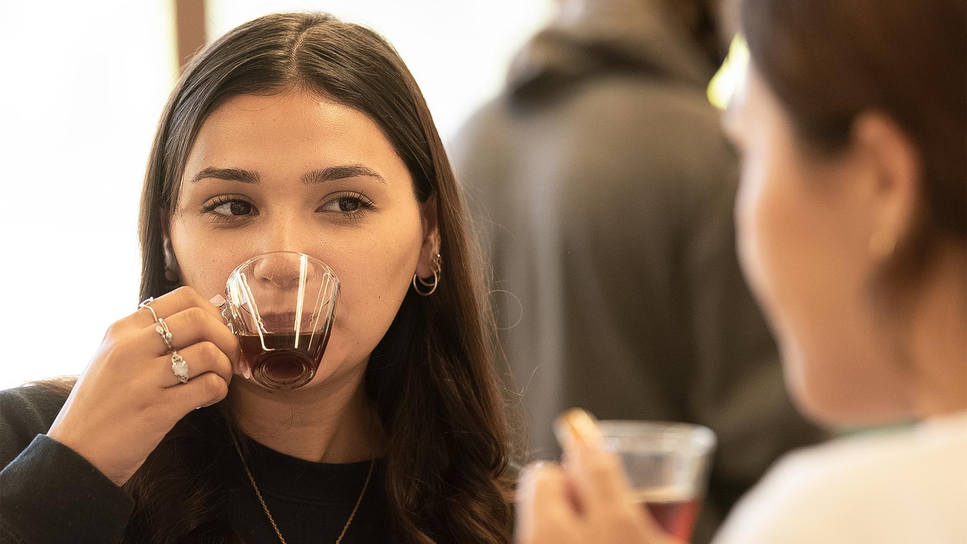 A coffee class student raises a small cup of coffee to her lips to taste as she looks at her lab partner