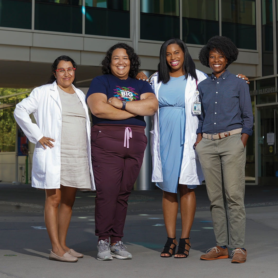 A group of four women stand together in front of a medical office building. Two wear lab coats and one wears scrubs. One has a stethoscope. All are smiling and looking directly at us.