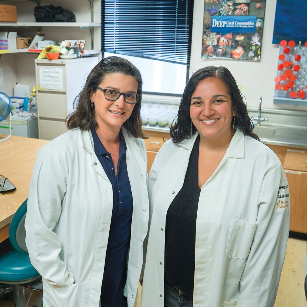 Two women scientists in lab coats stand together in a lab, facing us and smiling.