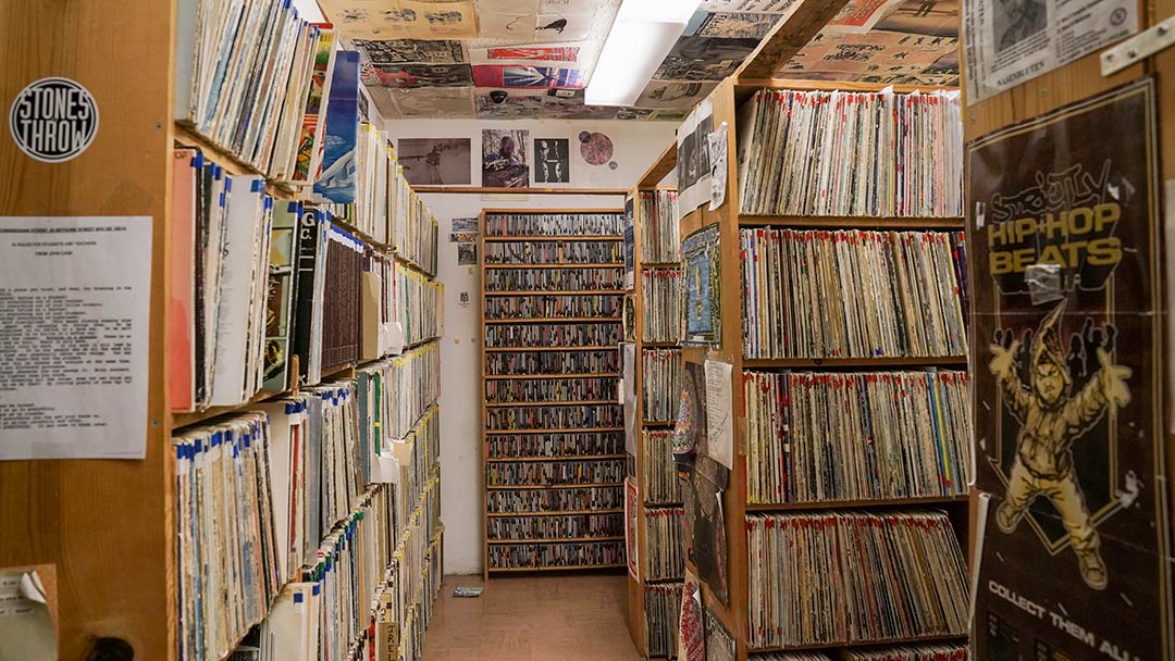 Photo of a small room with wall-to-wall, floor-to-ceiling racks of vinyl records, cassette tapes, CDs and music paraphernalia.