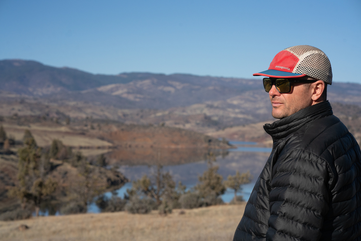 Robert Lusardi in black jacket and red cap looks above Iron Gate dam in 2023