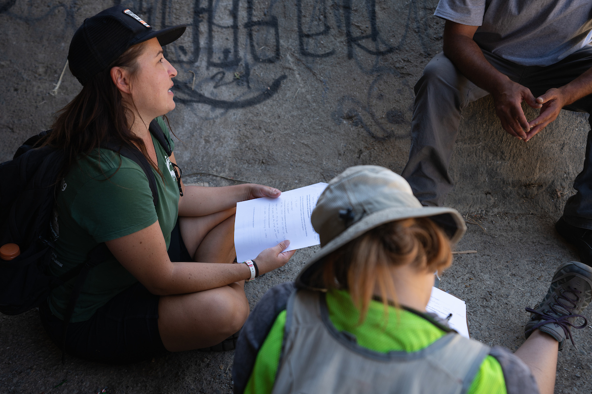 Costanza Rapini in cap hold clipboard sits in graffiti-covered tunnel talking to unseen person while woman in bucket hat and yellow vest sits next to her, head bent over clipboard