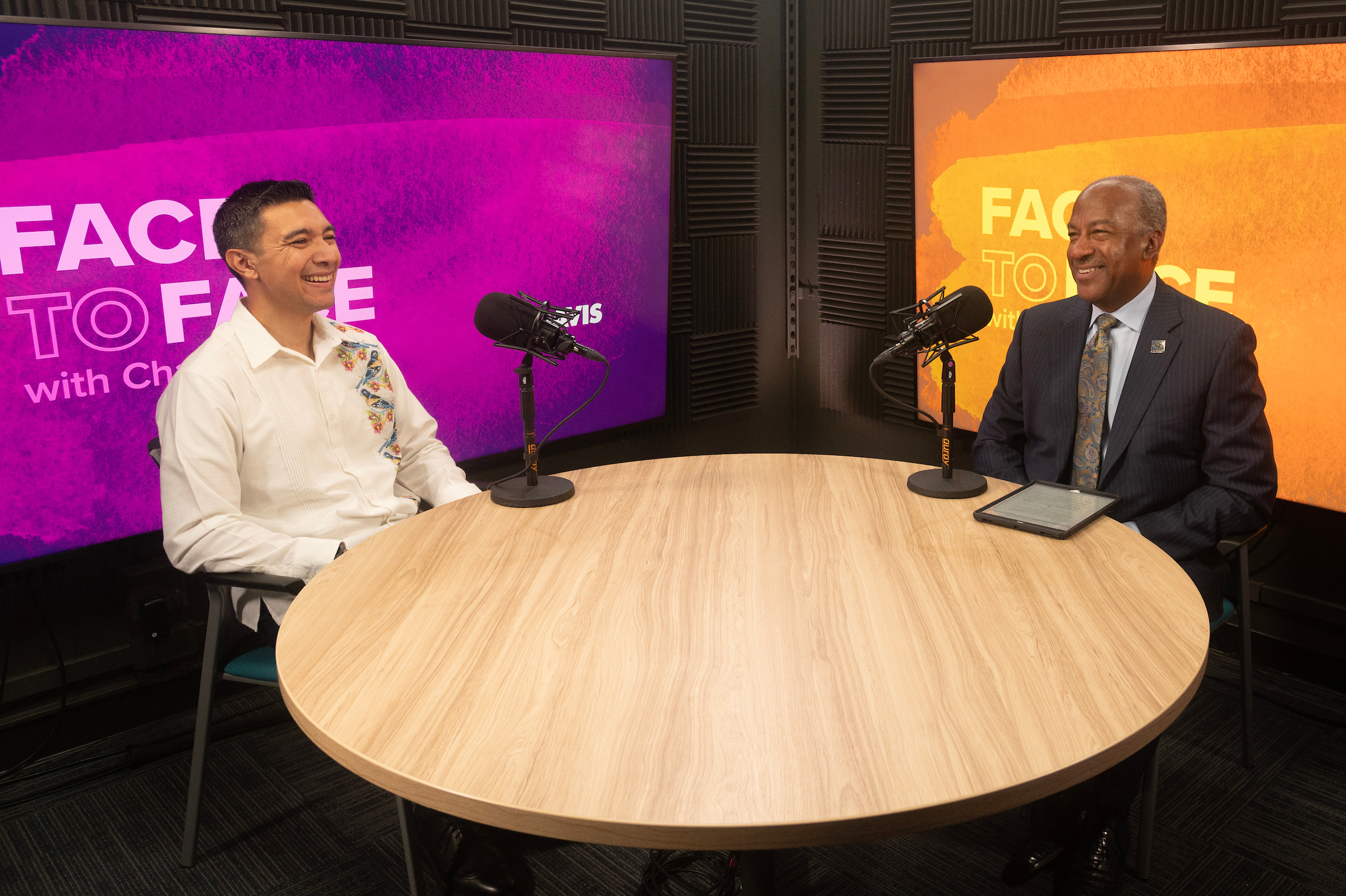 Two men sit across from each other at a round wooden table inside of a recording studio. The man on the left is wearing a white shirt with a bird pattern along its right side. On the right side, Chancellor Gary S. May wears a dark suit and listen to Ramírez's talking points.