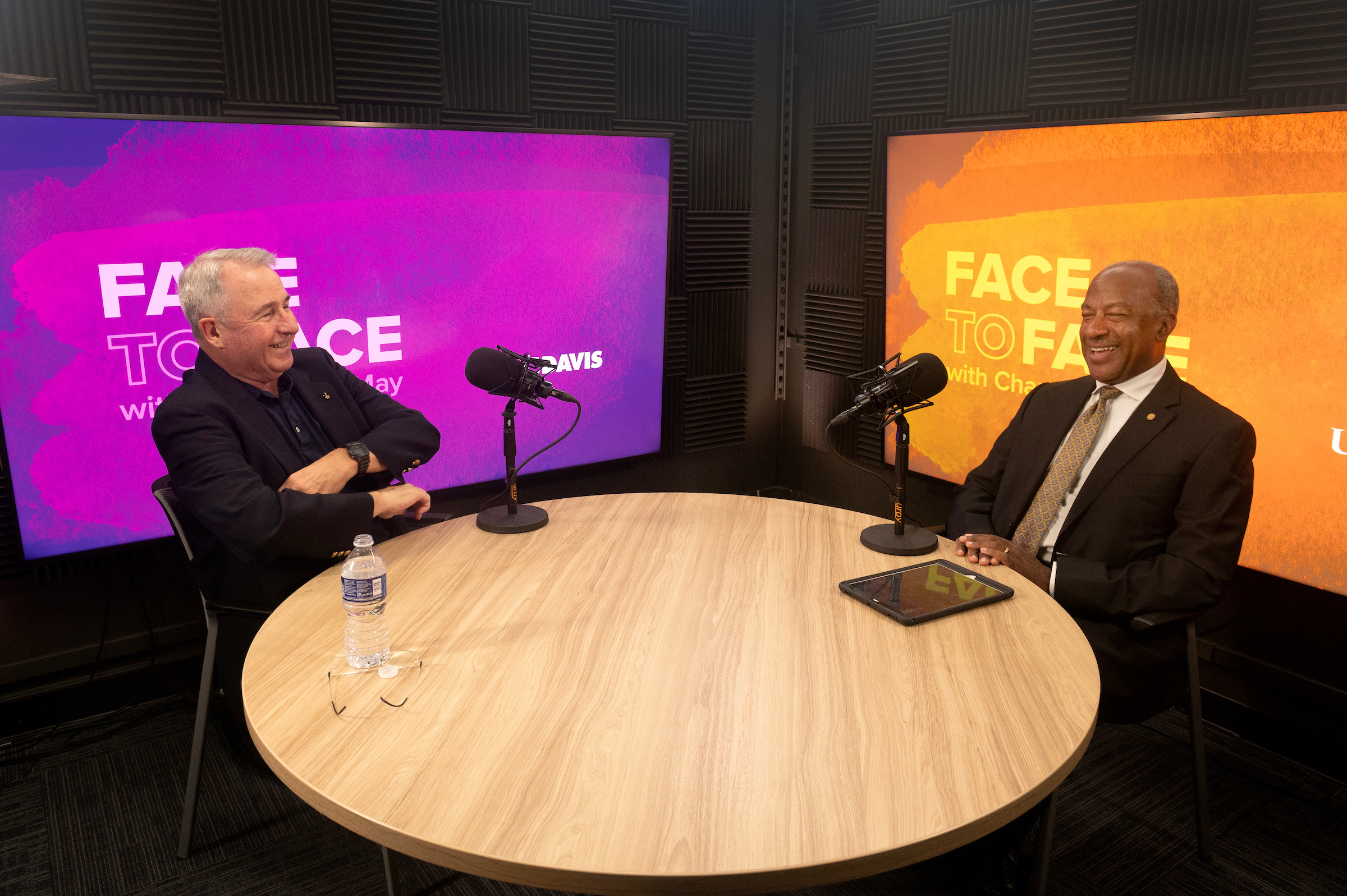 Two men sit across from each other at a round wooden table inside of a recording studio. The man on the left is wearing a dark suit jacket, has short white hair and has their arms crossed. On the right side, Chancellor Gary S. May wears a light brown suit and listen to Professor Steven Robinson's talking points.