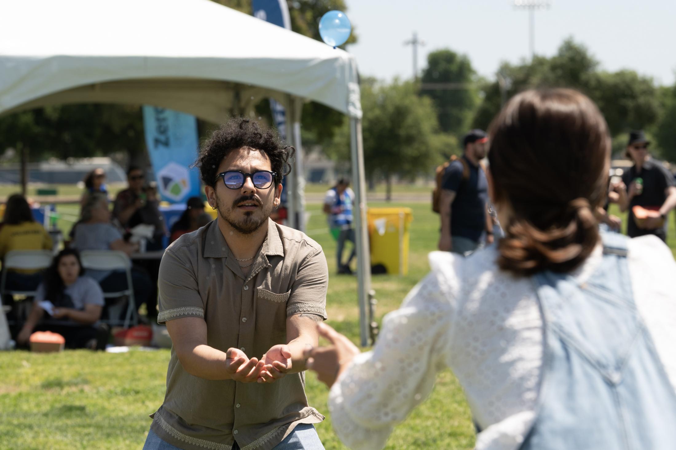 A person reaches forward with both hands to catch a water balloon during a campus event, while another person stands in front of them, ready to catch the ballon with their back to the camera. 