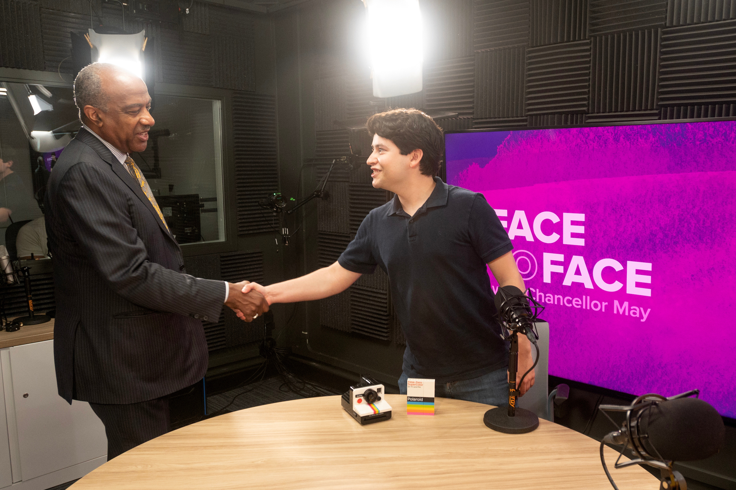 Inside of a recording studio, Chancellor Gary S. May, wearing a dark suit, is standing on the left and extending his hand for a shake with Marc Corfmat, standing to the Chancellor's right, wearing a dark polo shirt against a Face to Face logo backdrop. 