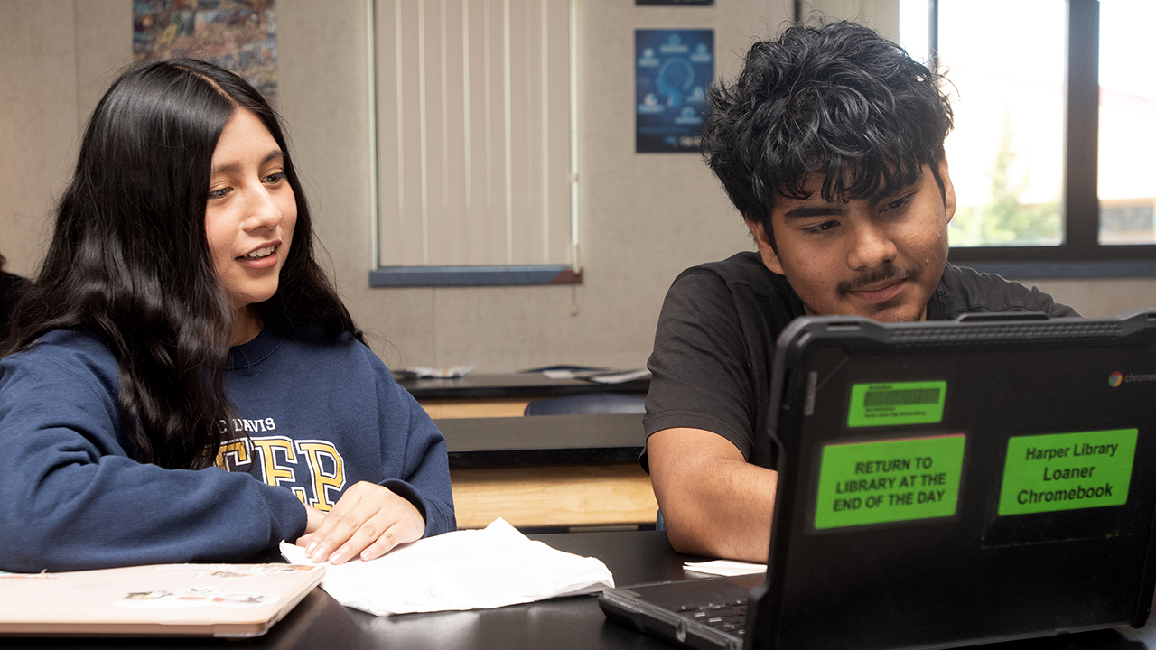 A woman sits at a table with a student working at a laptop.