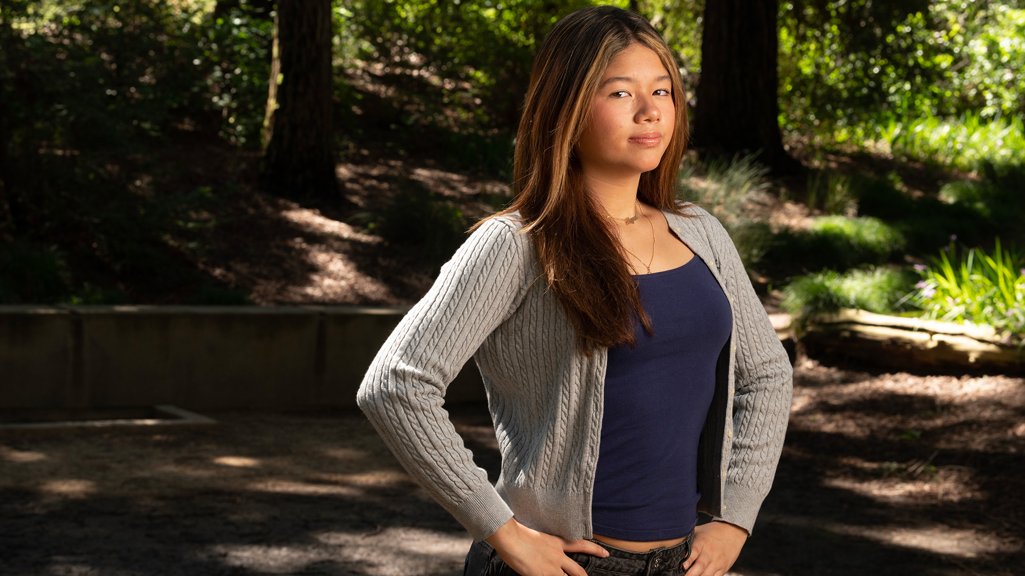 Young woman in gray cardigan and navy top standing in sunlit park, hands on hips, confident