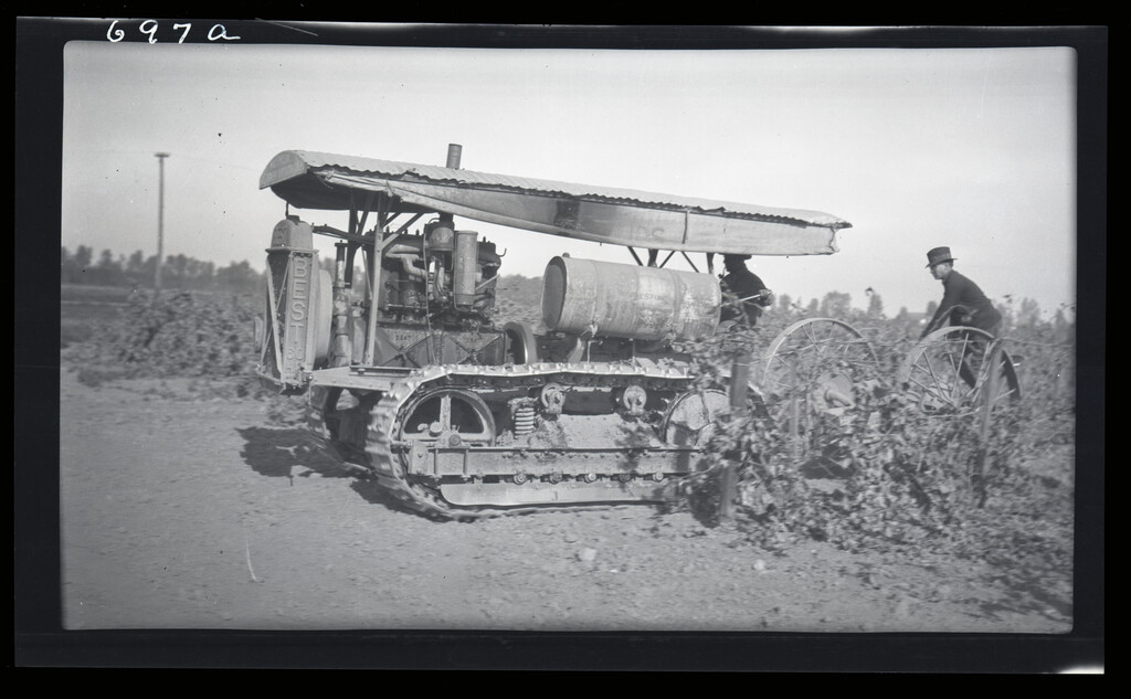 Two workers using a vintage tractor in a field, with crops in the foreground.