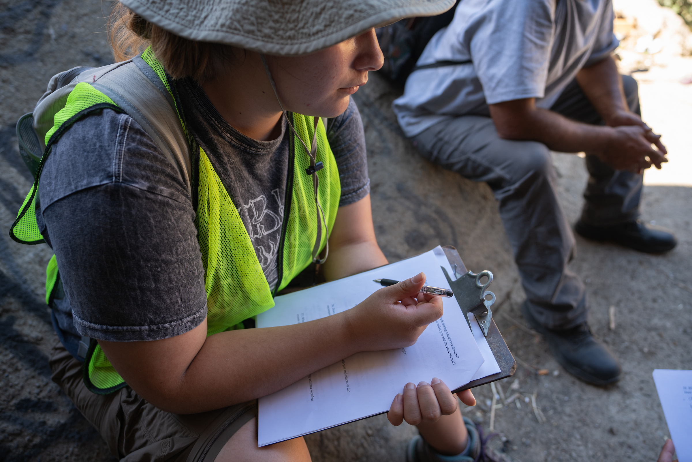 woman in hat and bright yellow vest holds clipboard with white paper. The torso and legs of a man sitting next to her is visible.