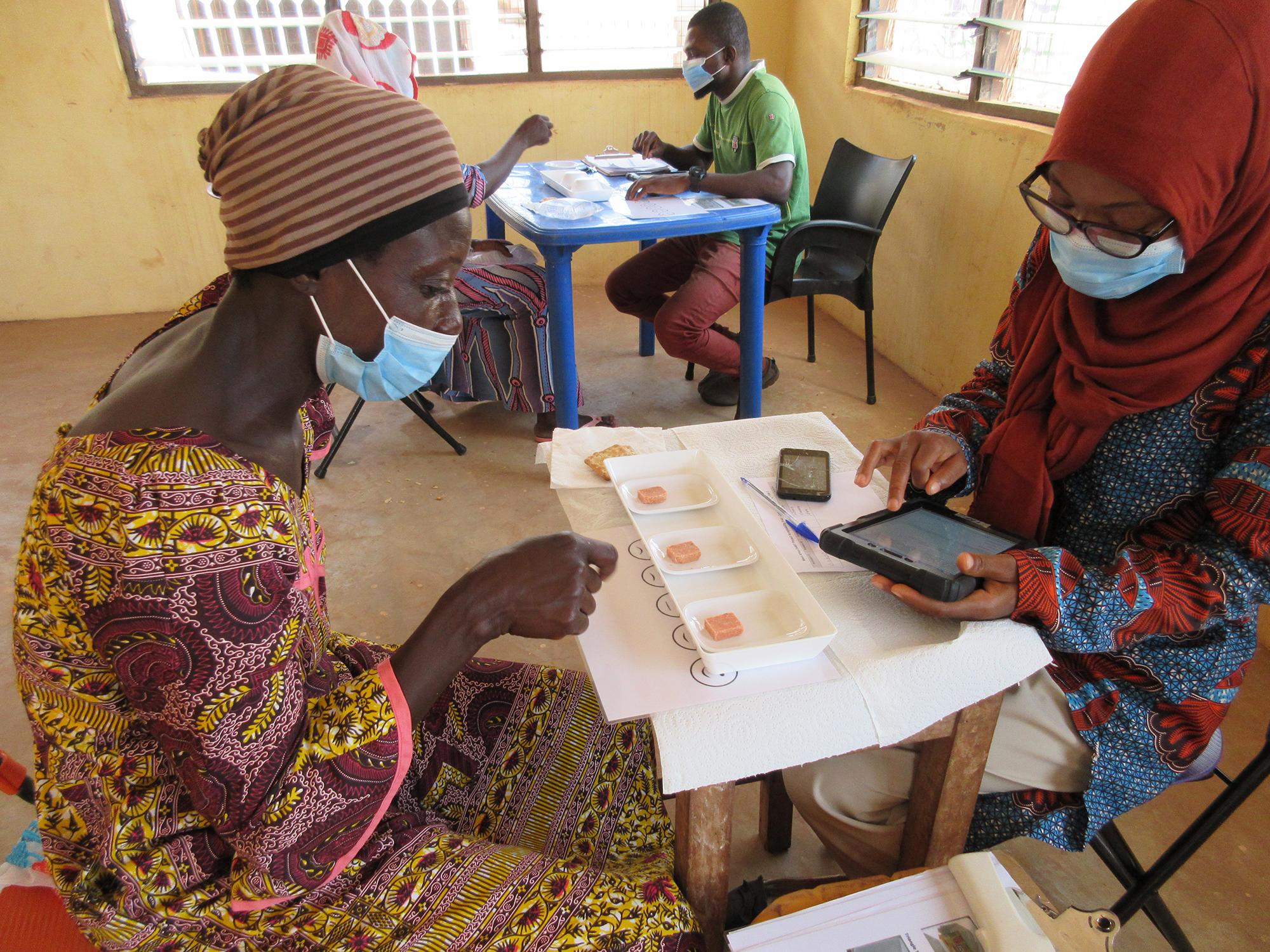 Two African women sit at a small table. The woman at left is looking at a white tray with yellow cubes in three wells. The woman at left takes notes on a tablet device. 