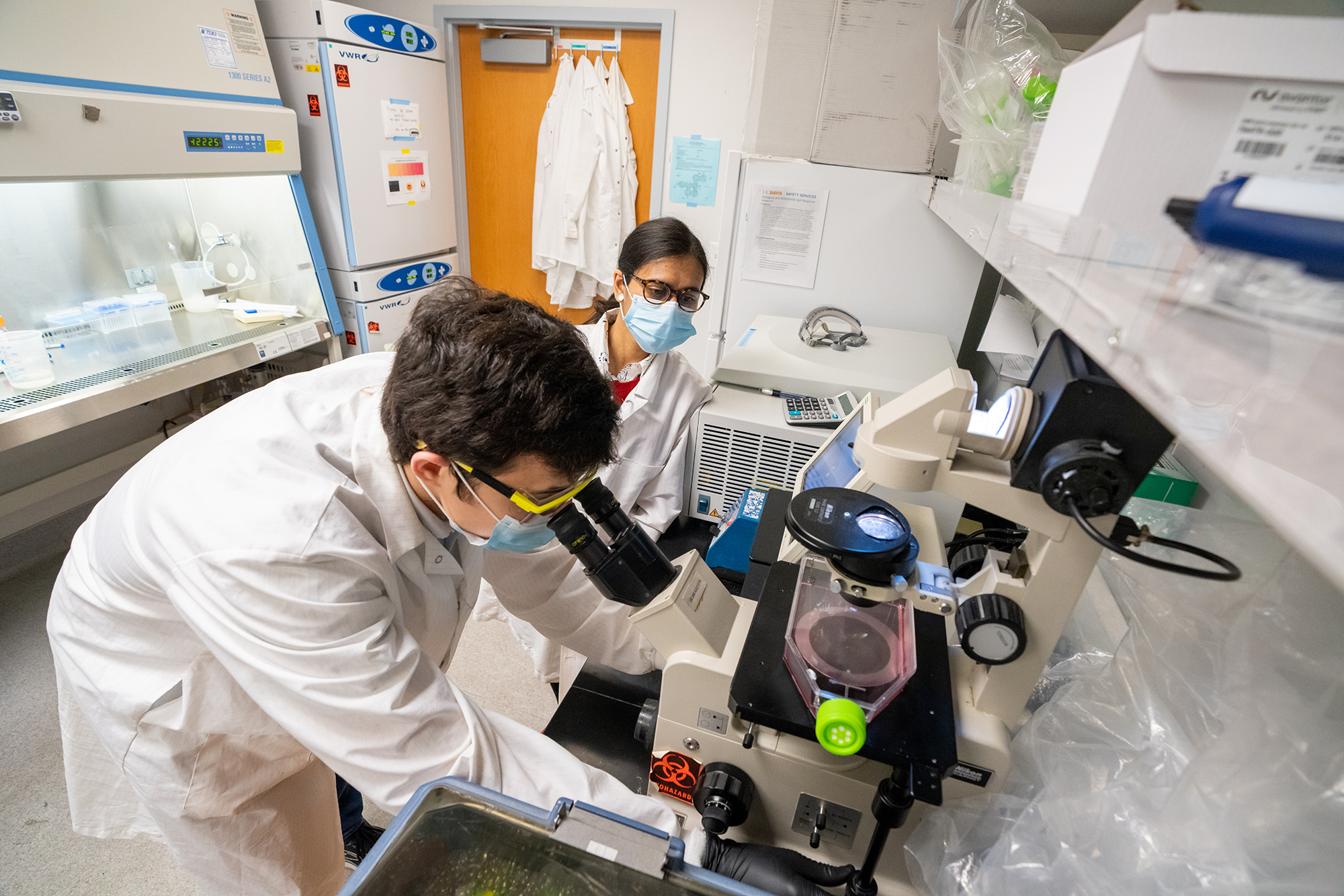 In foreground, a young man in a white labcoat, facemask and safety glasses leans over a lab bench. In background a woman with long black hair, safety glasses and face mask watches. 