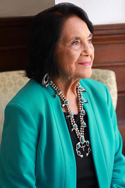 Woman seated, teal blazer and layered silver necklaces, gazing upward with a soft smile