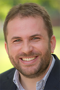 Smiling bearded man headshot in blazer, blurred green outdoor background