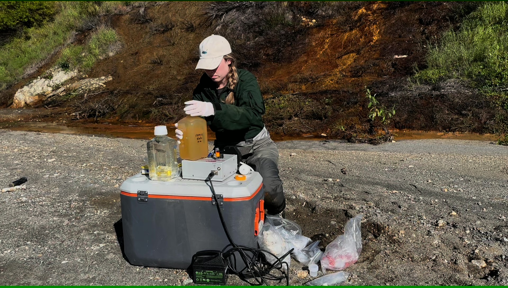 Scientist Taylor Evinger in white hat with sampling gear on rocky shore, photo