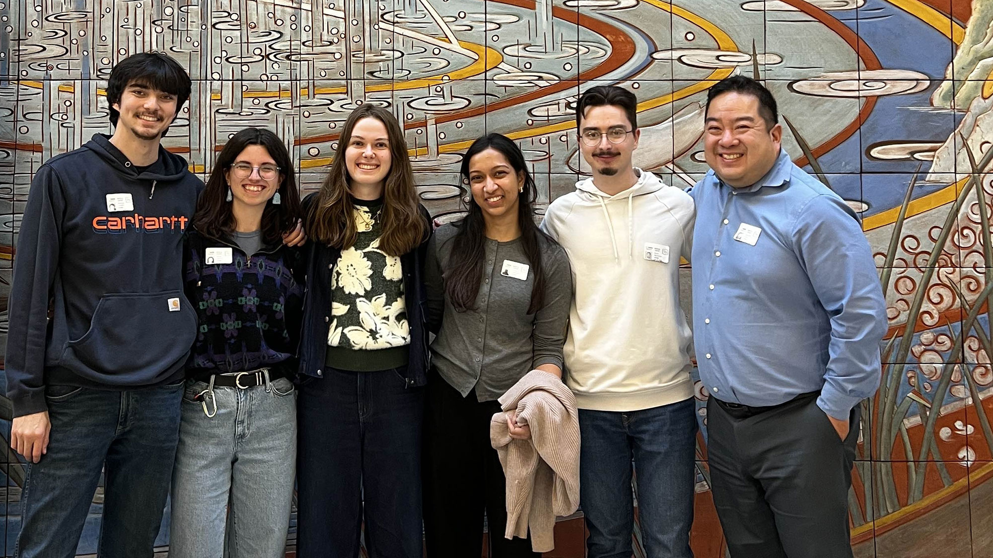Group of six smiling adults in front of colorful mosaic mural