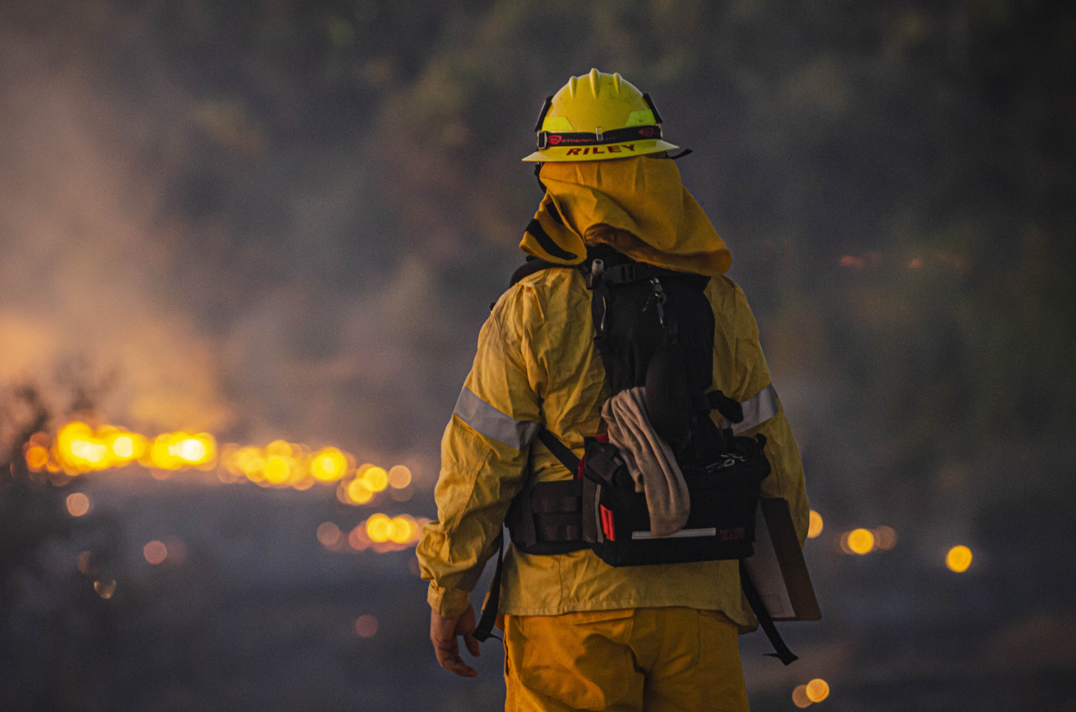 Firefighter in bright yellow helmet, jacket and pants seen with back to camera facing dark, smoky landscape with burning lines of fire on ground. 