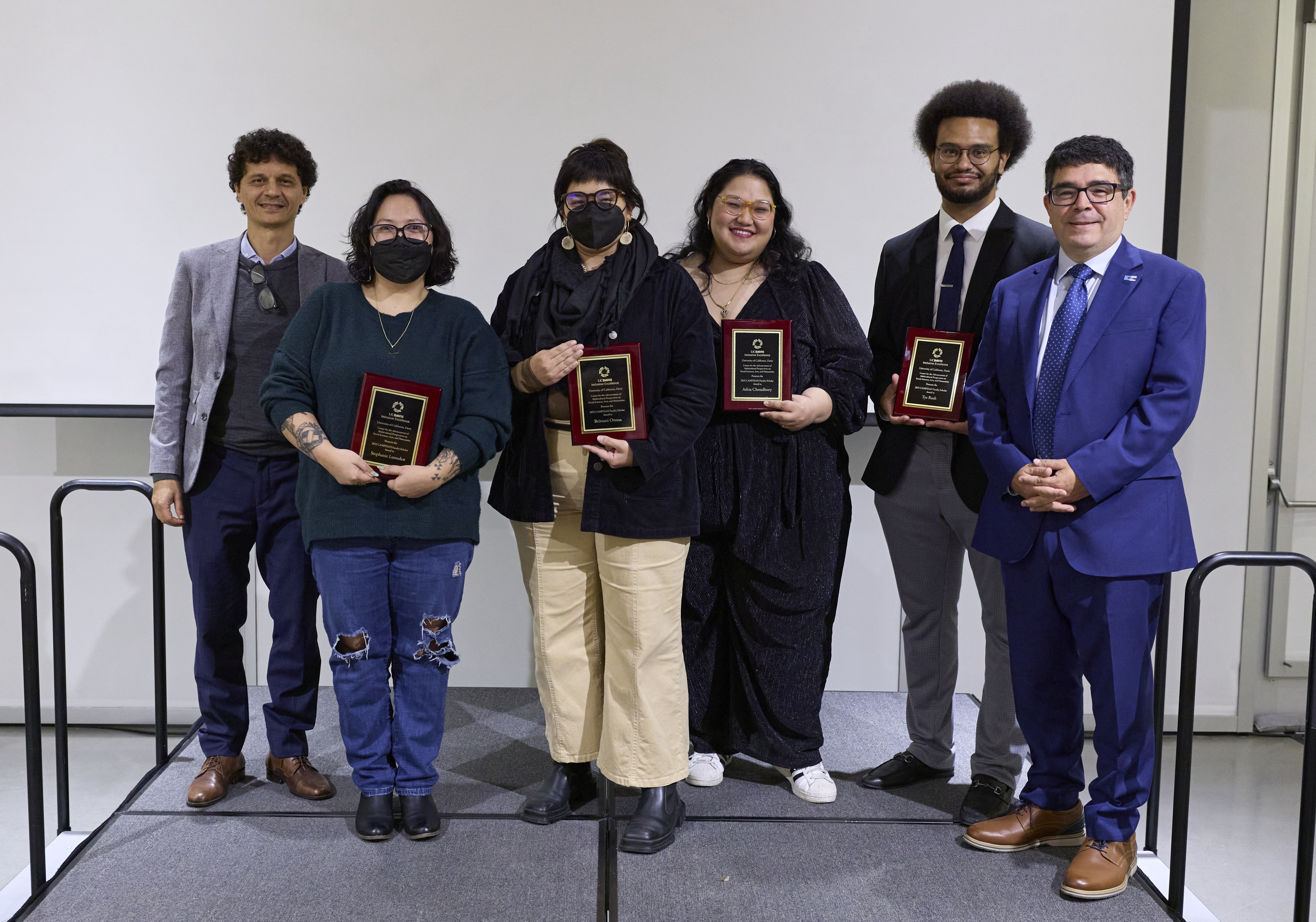 Six academics in professional dress stand in a row facing the camera on a stage in front of a blank projection screen. Several of the adults featured are holding plaques in their honor. 