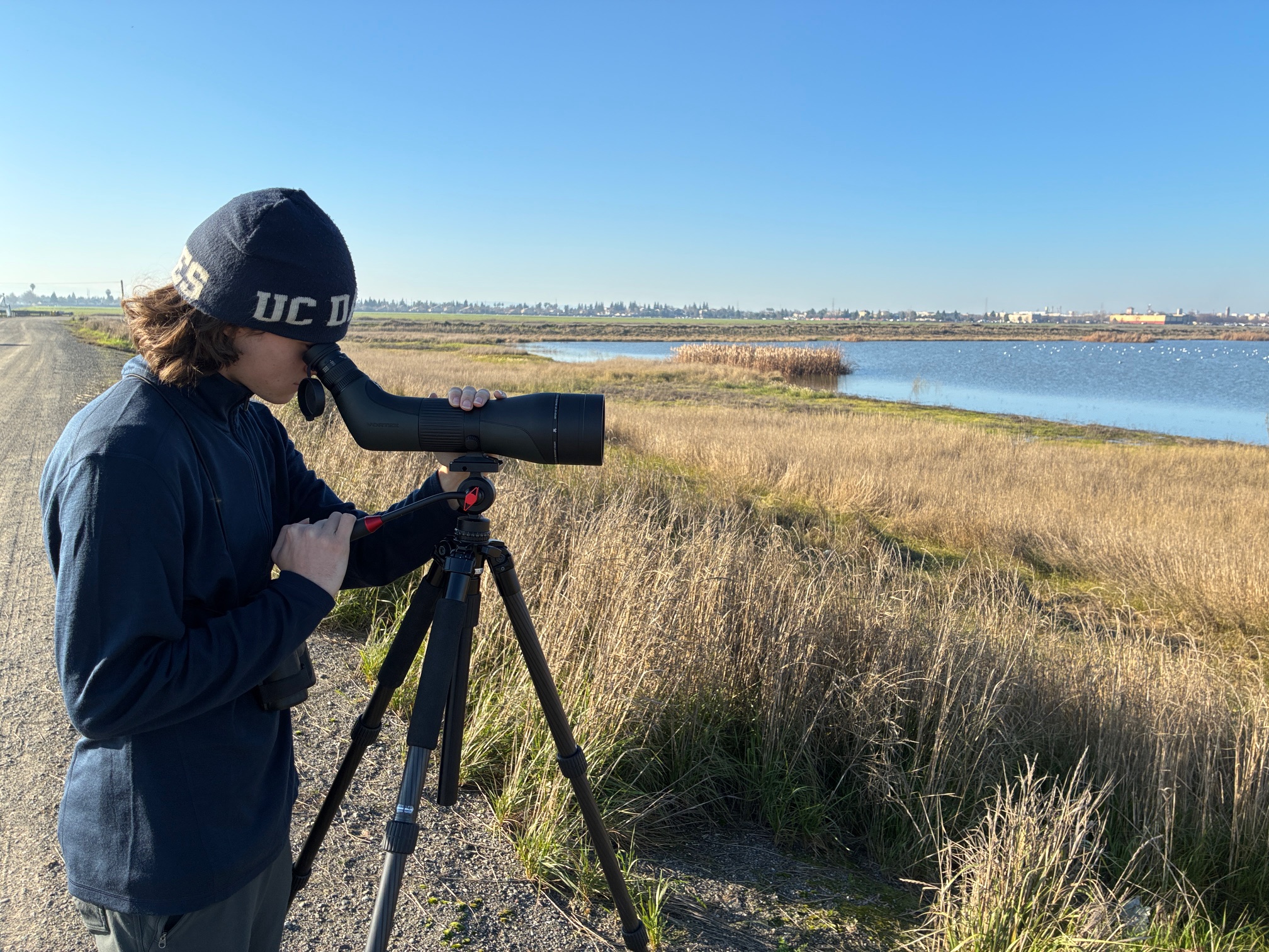 Julian Johnson looks through spotting scope toward waterbirds on pond, just past grassy floodplain