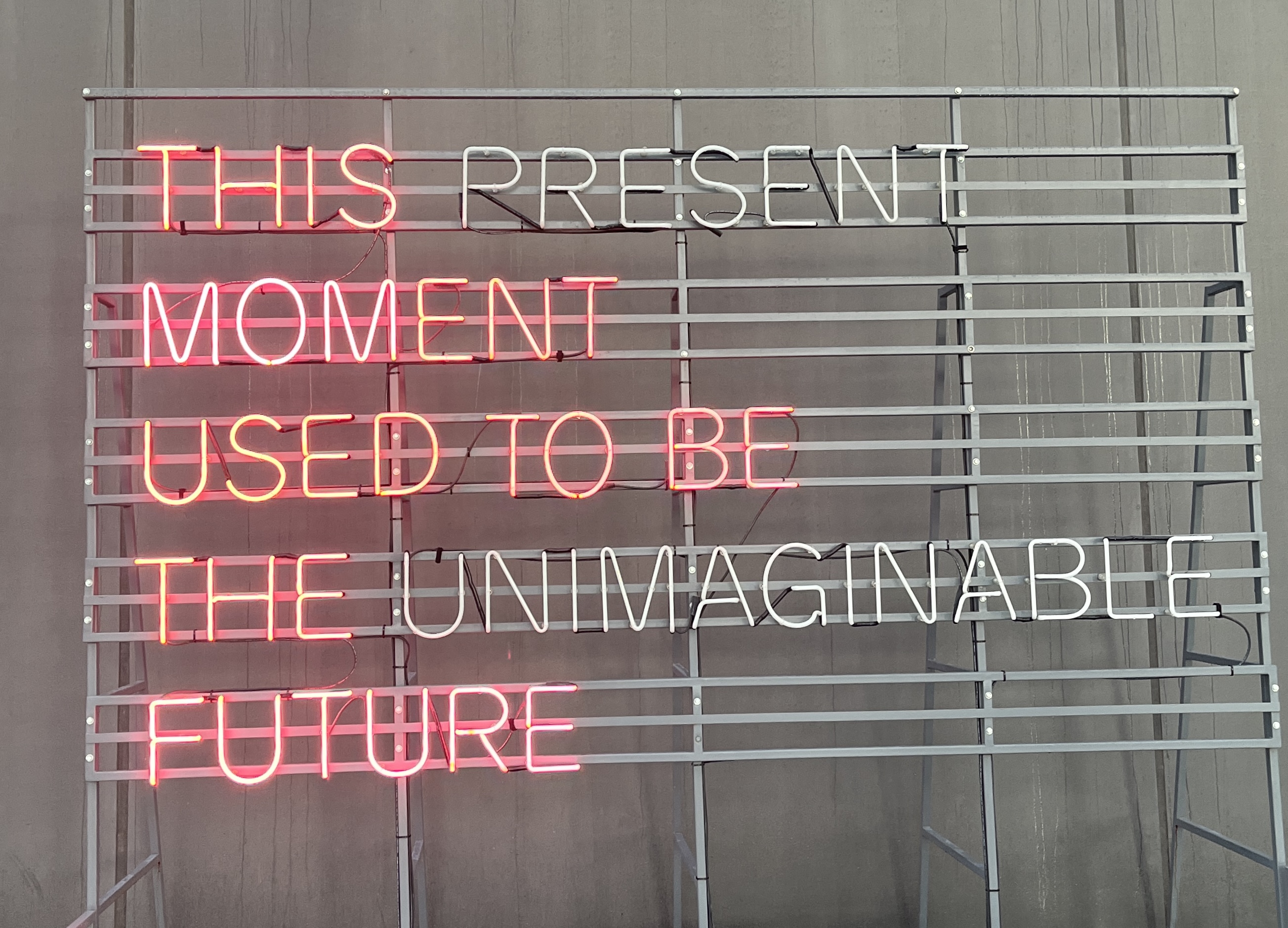 Neon sign with pink letters spelling out “This present moment used to be the unimaginable future.” 