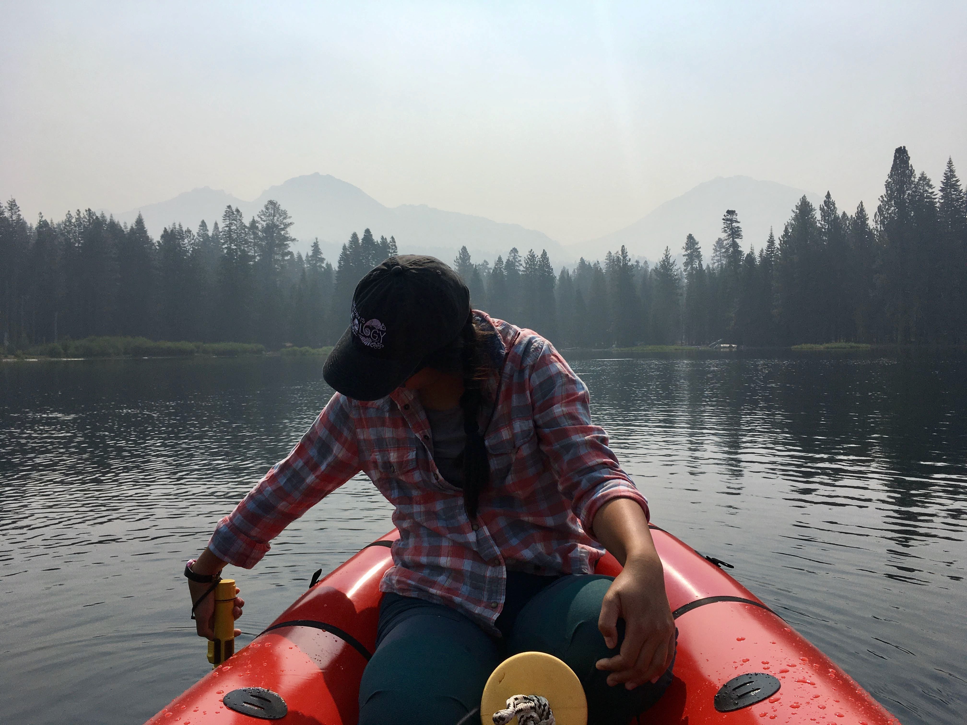 Ecologist MJ Farrugia on Manzanita Lake in a red kayak leans over to sample water under smoky skies.