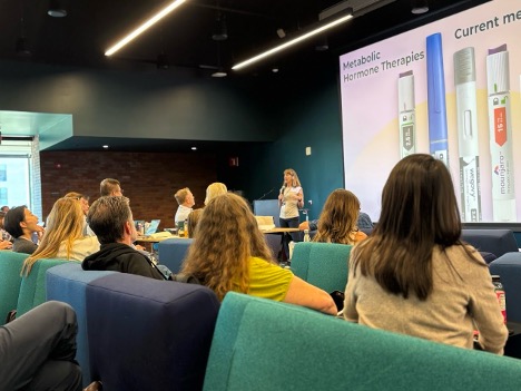 A woman holding a microphone and presenting in front of a mixed audience. A slide behind her with a picture of metabolic hormone therapies.