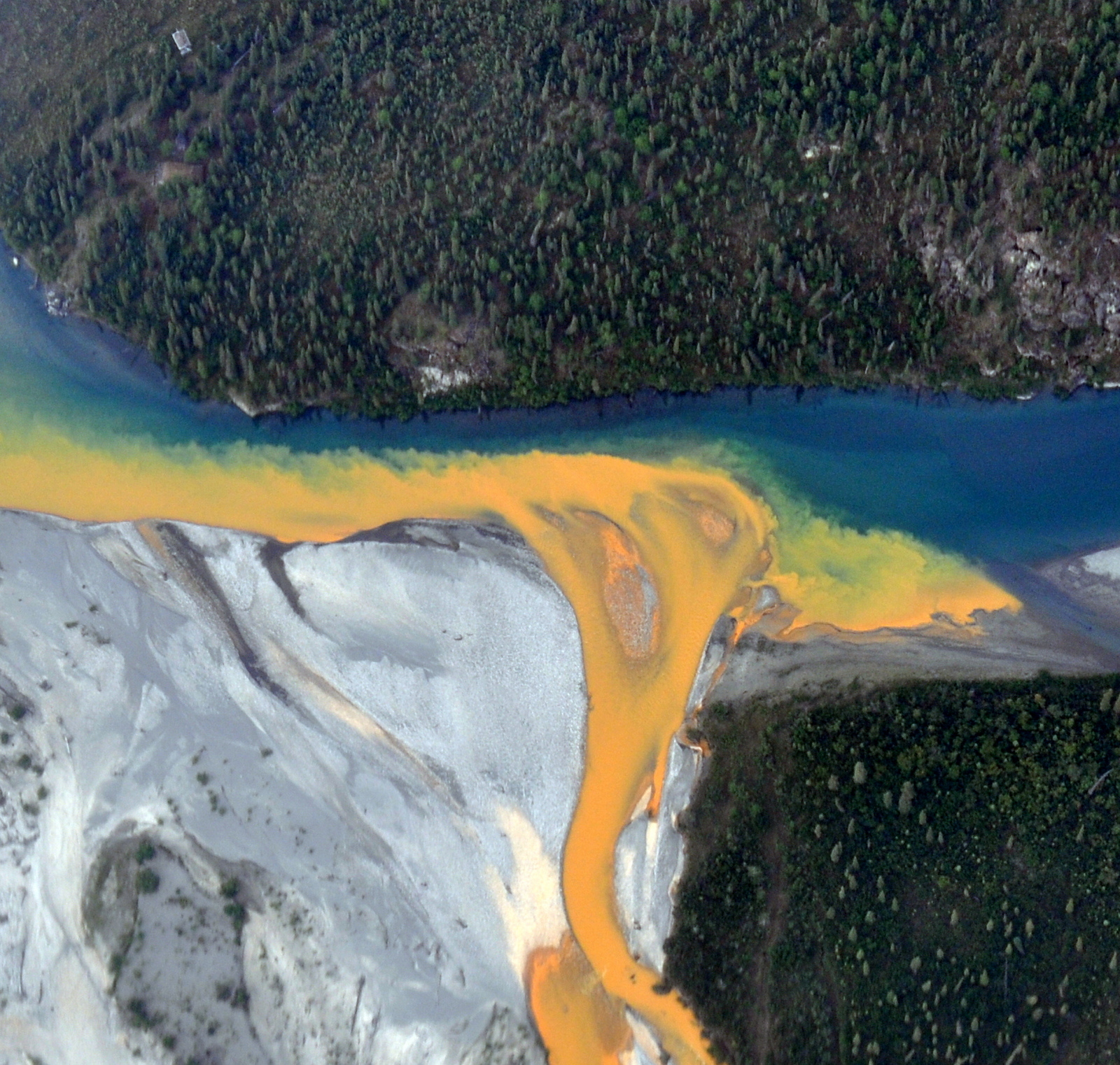 Ribbon of bright orange melds with greenish blue water in aerial view of rusting river in Alaska