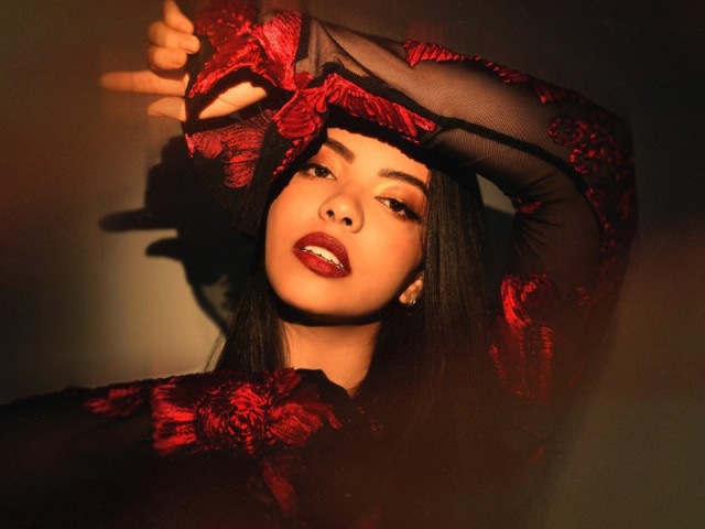 Portrait photo of sultry woman in red lace gloves and headpiece, hand to forehead, warm lighting