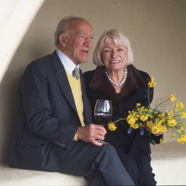 Older couple seated, smiling; man holds wine glass, woman holds yellow bouquet.