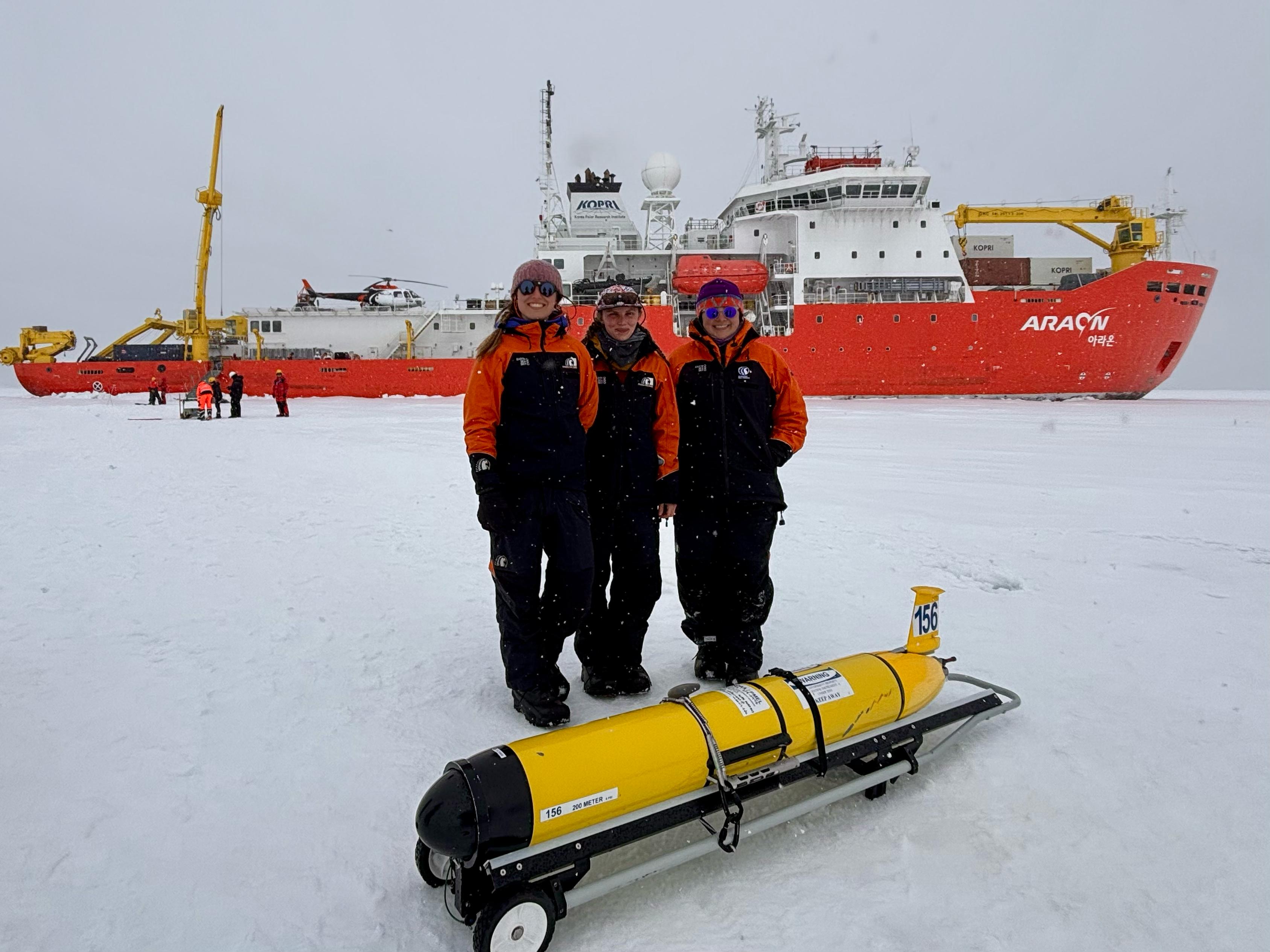 Three scientists in coats stand next to yellow robotic glider set down on ice and snow. Bright red and white icebreaker research vessel Aaron is behind them