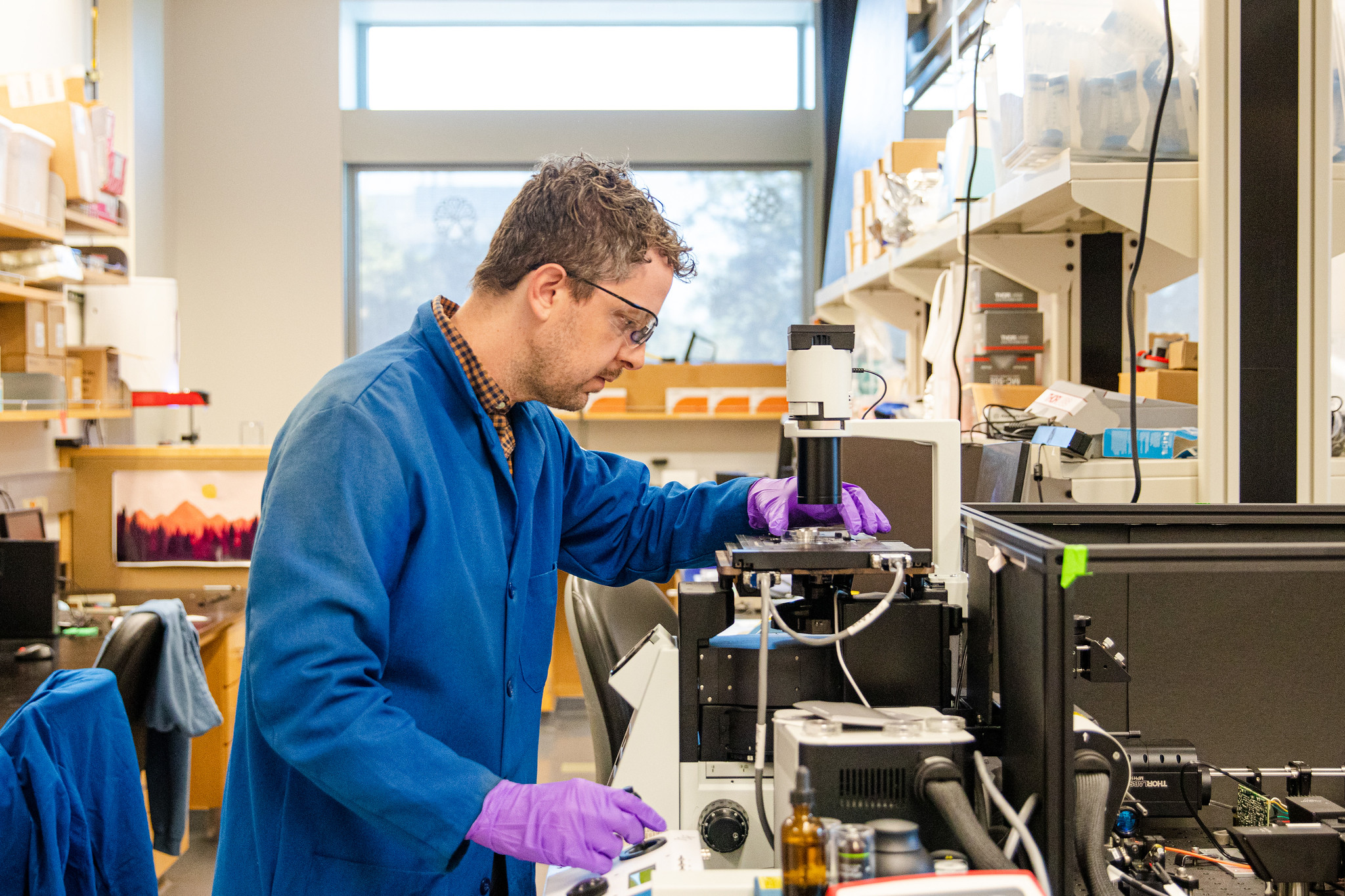 Randy Carney in blue lab coat and purple gloves using a microscope in a lab