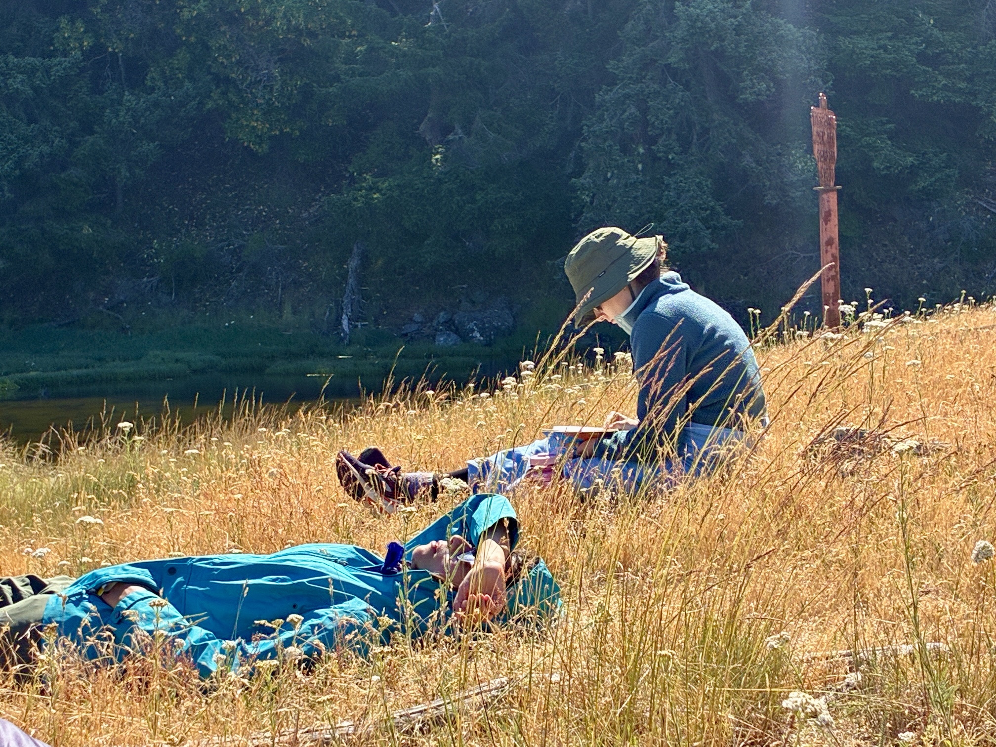 Two people relaxing in tall golden grass by a river, one seated with a sun hat