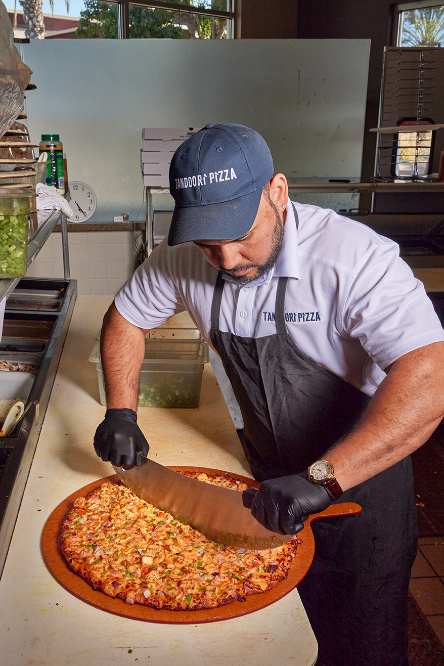 A man wearing a cap and gloves slices a pizza at a restaurant kitchen counter.