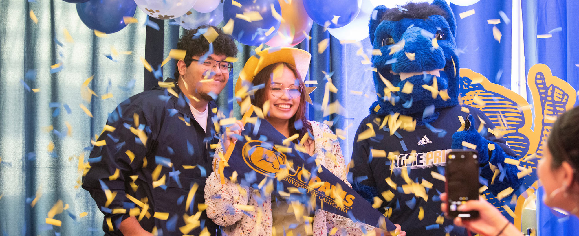 Two prospective UC Davis students pose with Gunrock the Blue Horse Mascot of the university while confetti is thrown in the air.