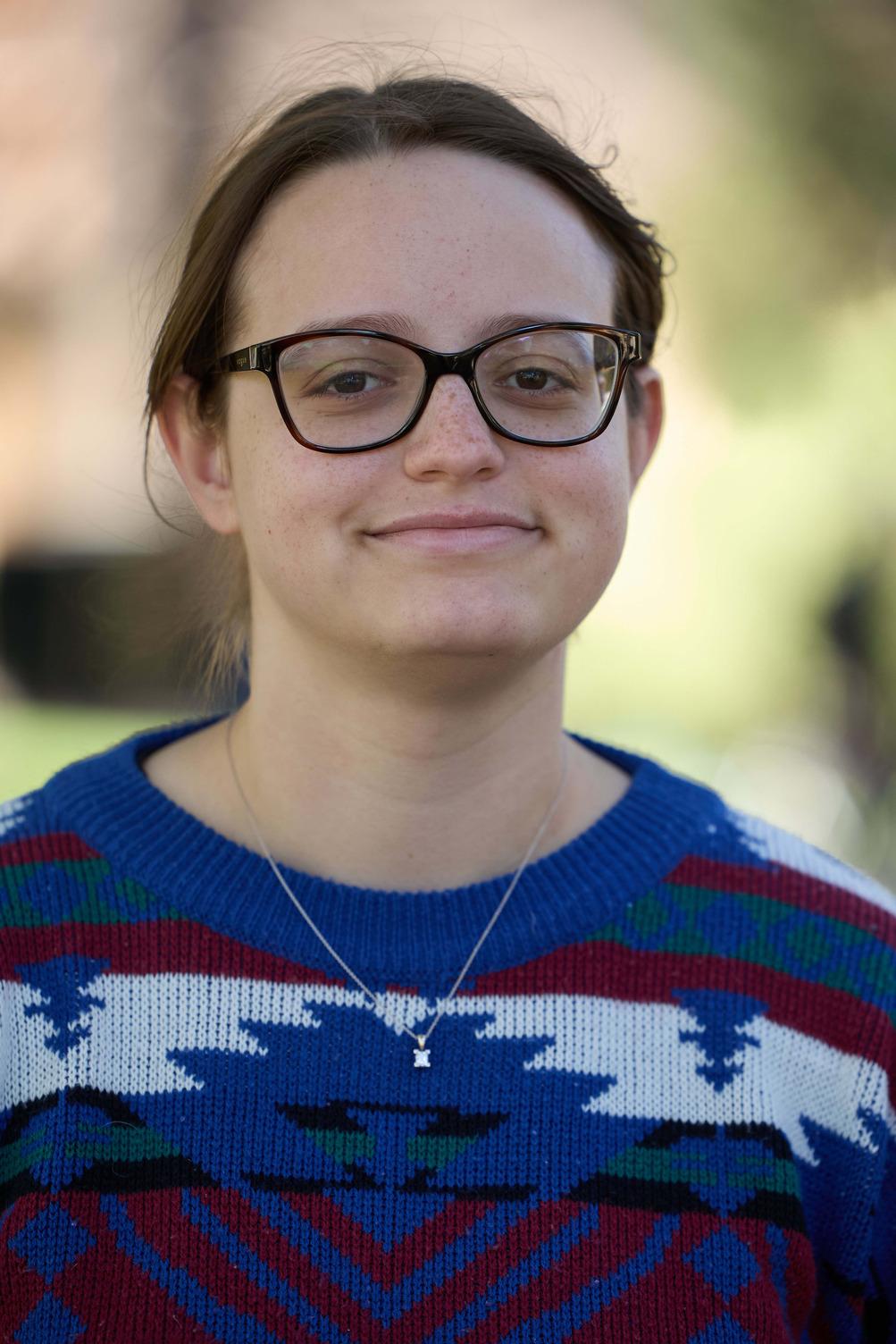 One student in a cobalt blue, dark red and white patterned sweater poses in front of a blurred outdoor background. 