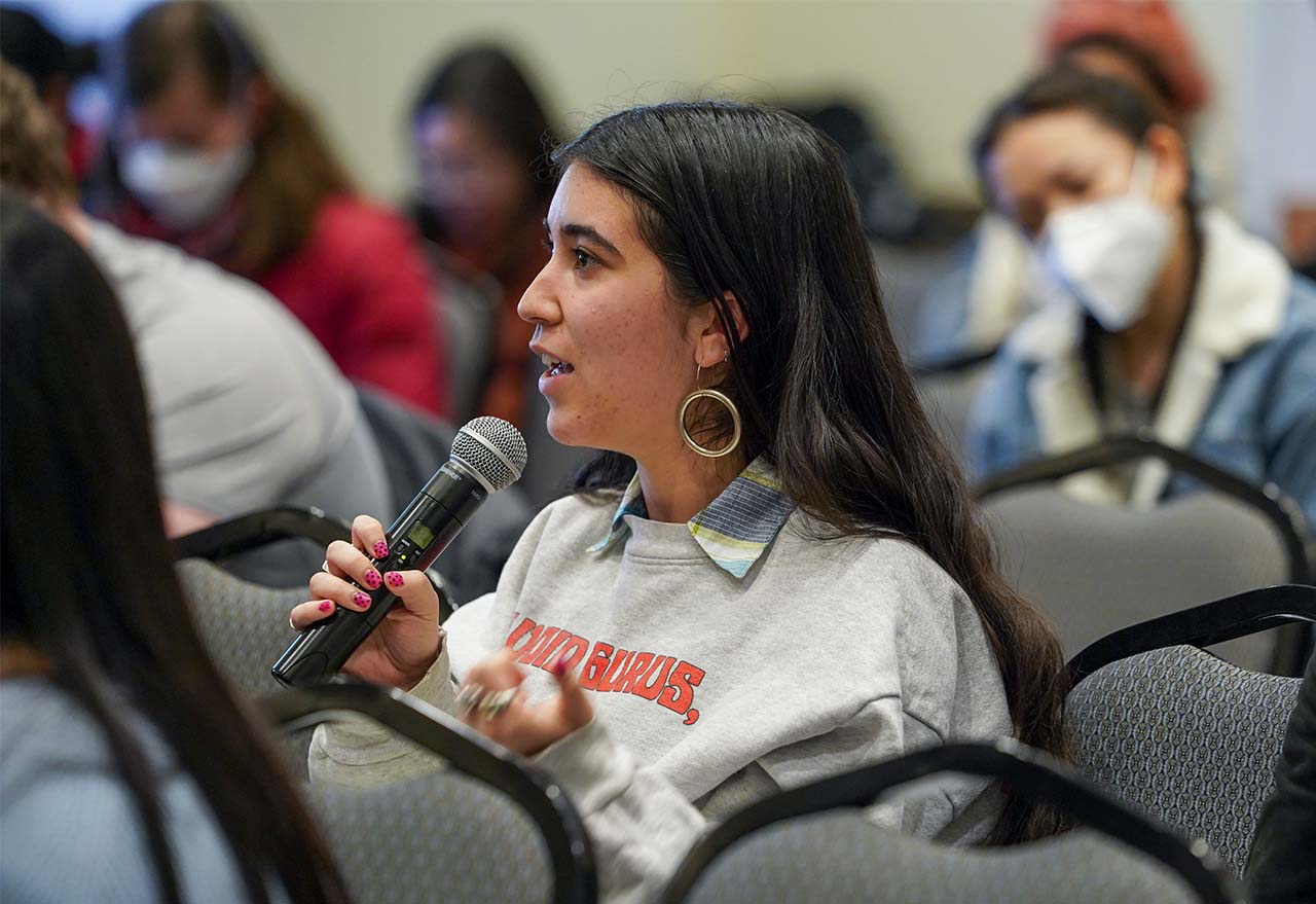 A woman wearing a light-colored sweater speaks into a microphone while seated at a town hall.