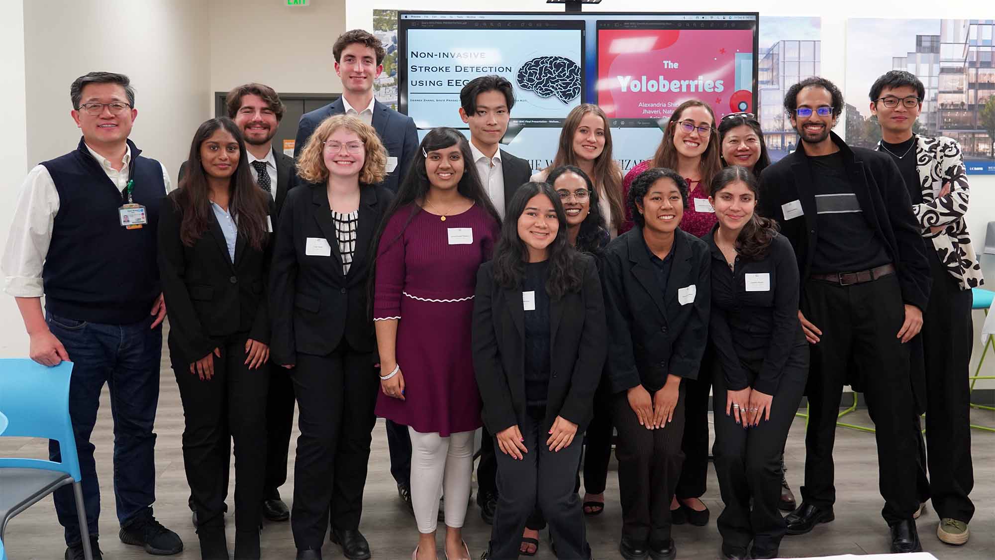 Group of 12 students, two faculty members and one peer mentor posing in front of a screen. The screen has images of presentations on them.