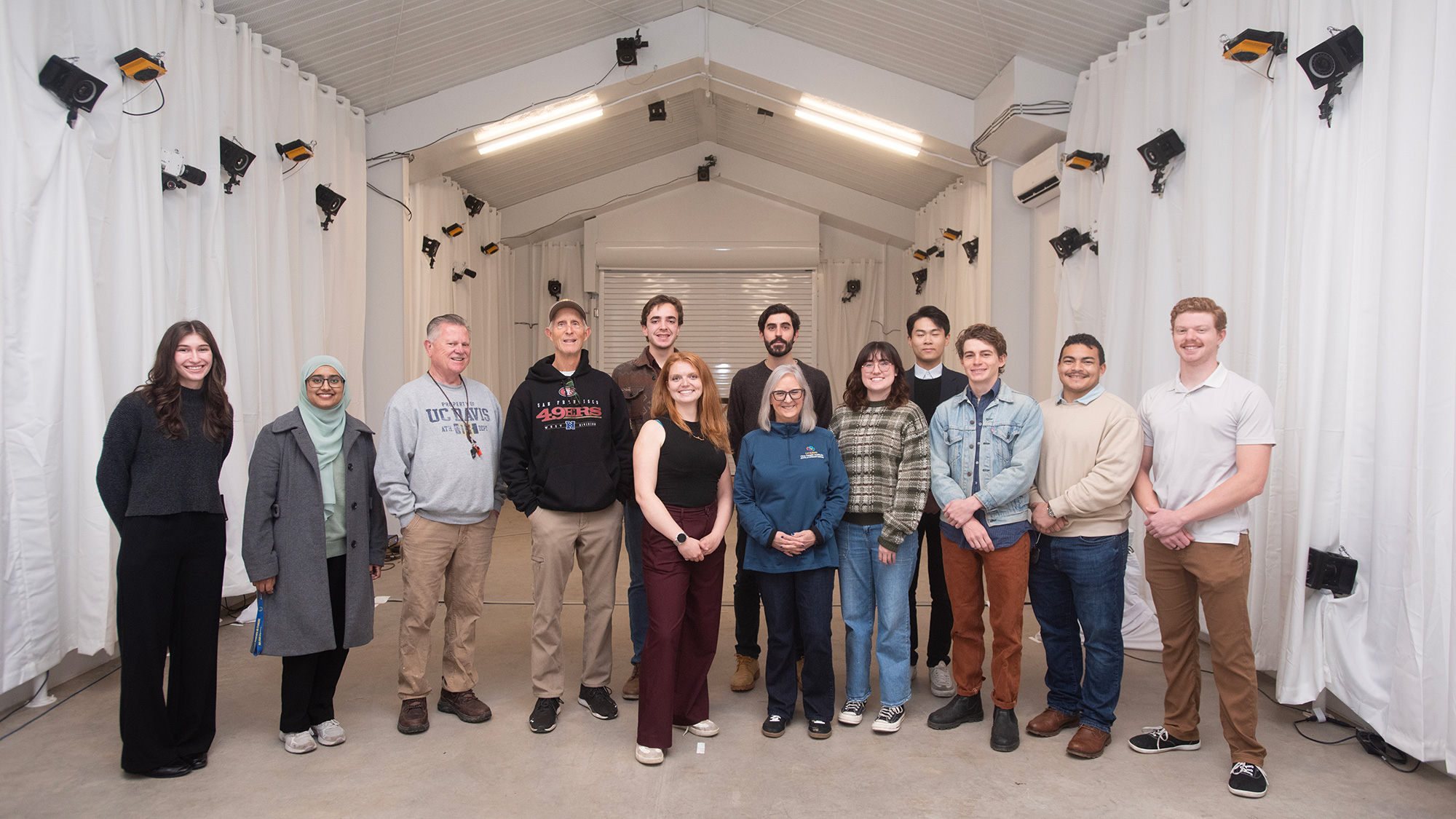 A diverse group of twelve people poses together in a well-lit indoor space with white curtains.