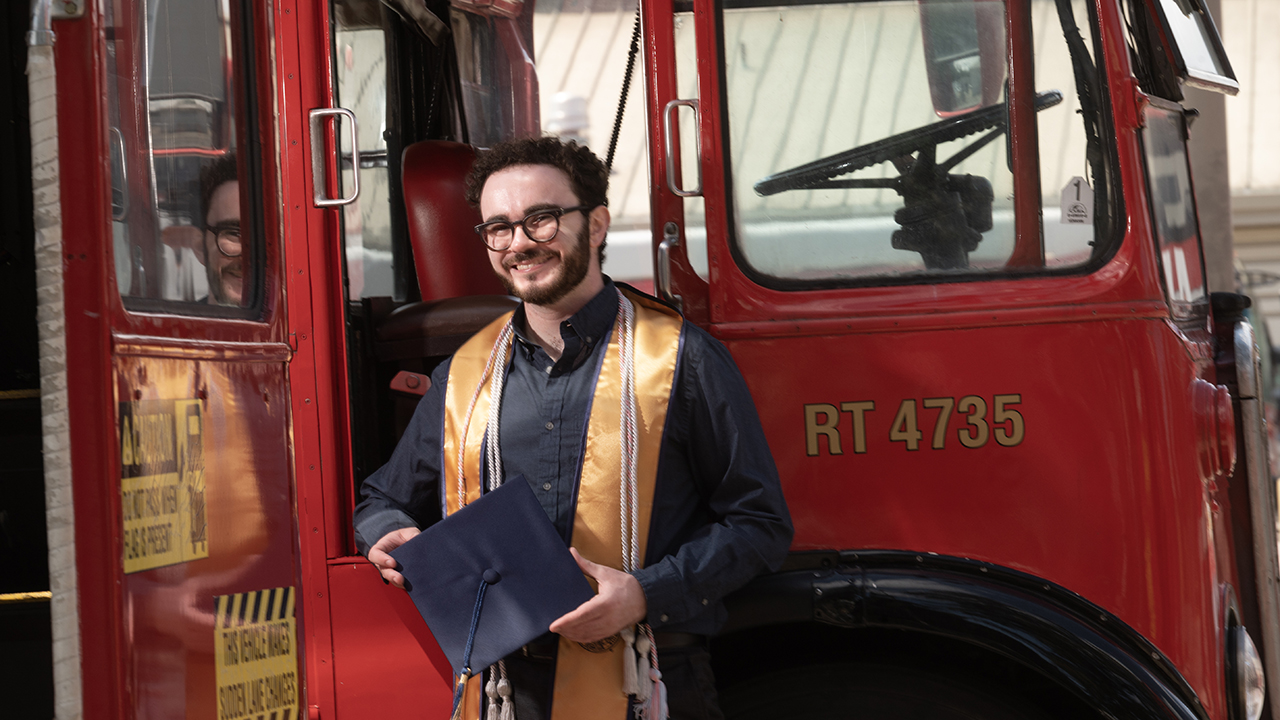 Smiling young man in graduation gown and gold stole holding cap and standing in front of a red vintage bus, smiling beside red vintage bus.