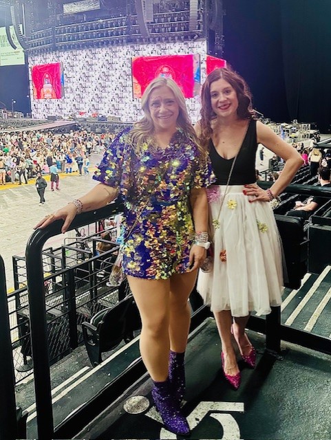 Two smiling women in cowboy hats and colorful dresses standing by railing at indoor concert