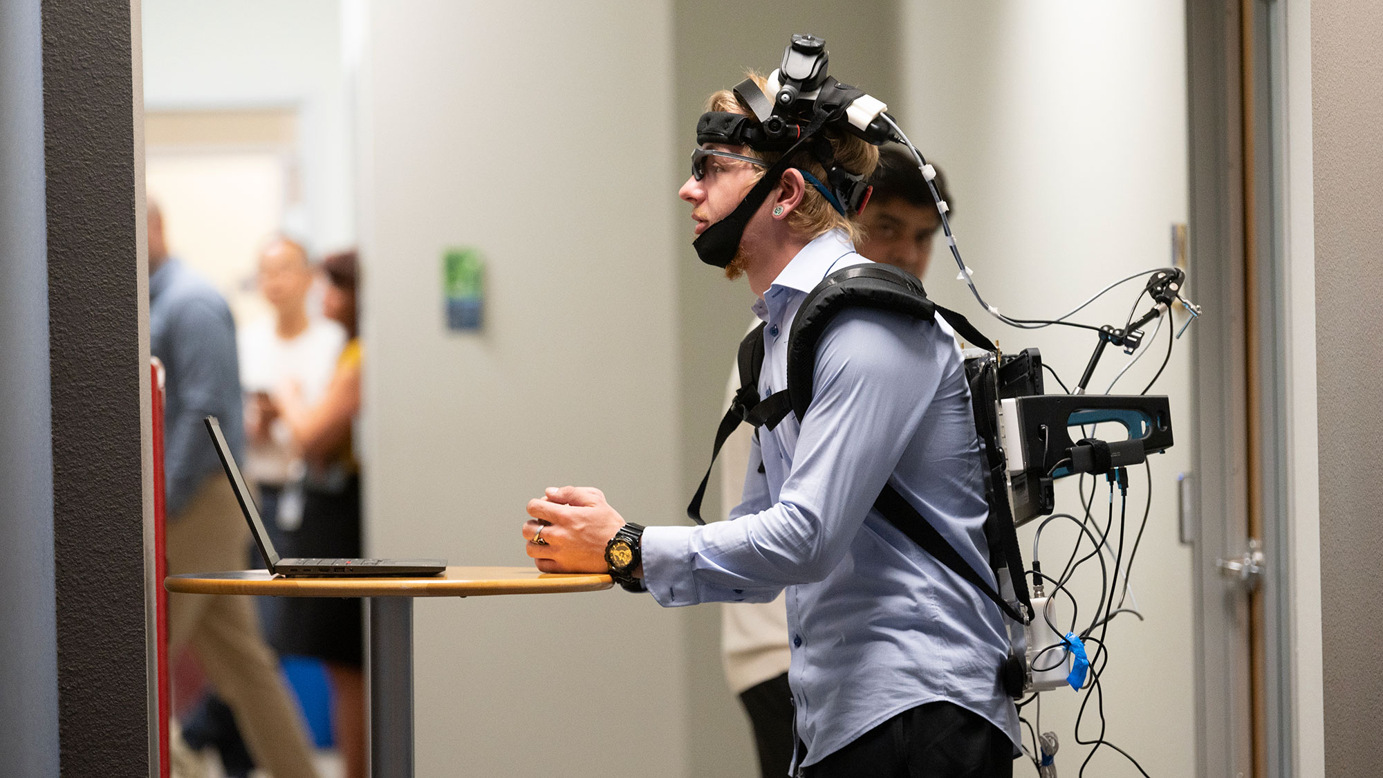 Person wearing head-mounted tracking gear and backpack, leaning on a table in a hallway.