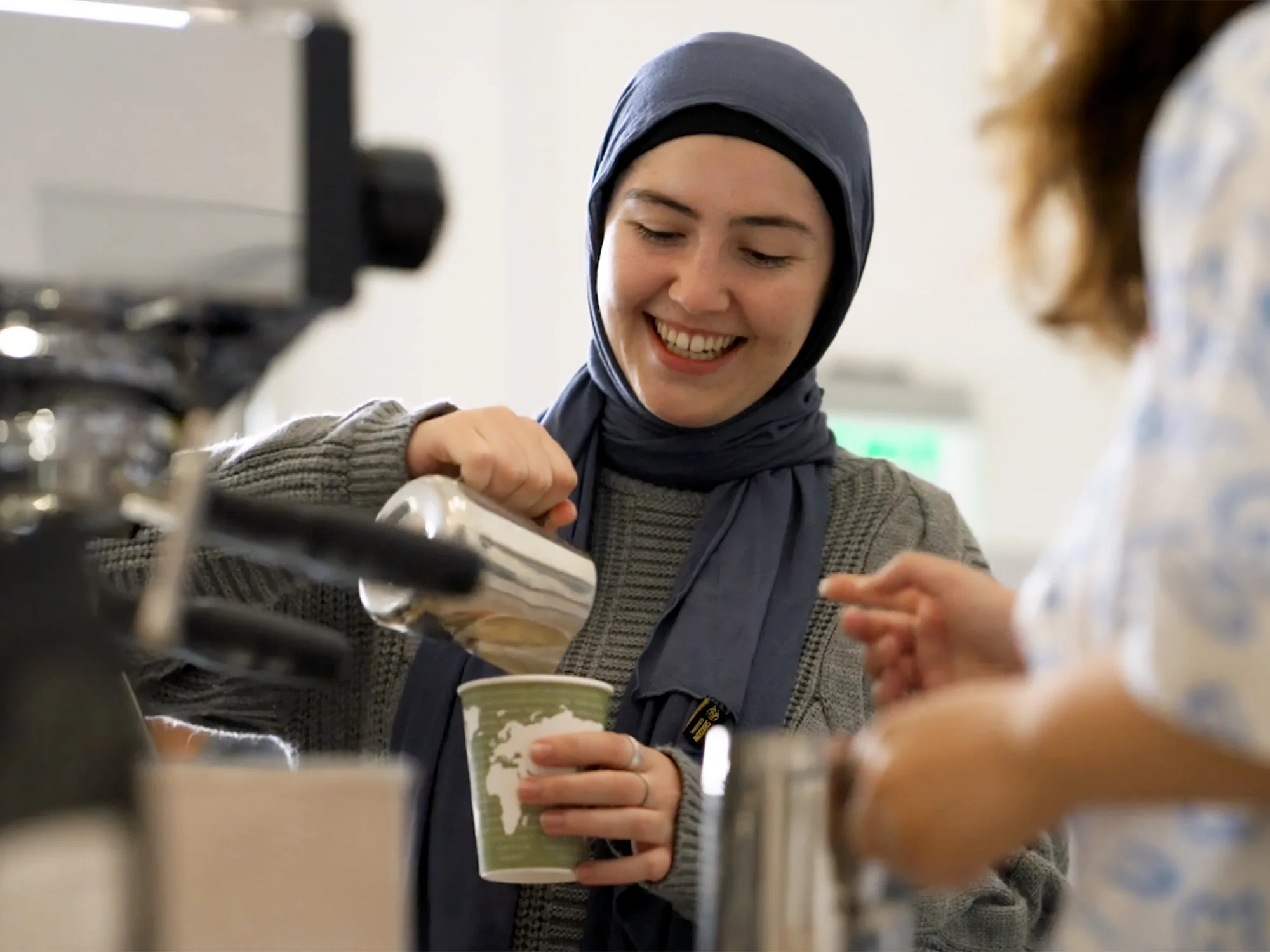 A smiling student in a hijab pours foam into paper cup with a green and white printed design of the Earth's continents.