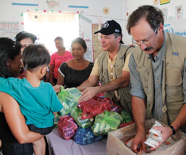 Two men wearing khaki vests pass out supplies to a mother and child.