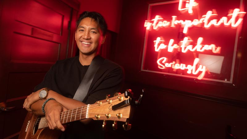 Photo of a man sitting and holding a guitar. He looks at the camera and smiles.