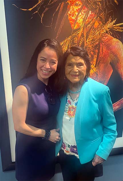 Two smiling women, younger in dark dress and older in teal jacket, colorful artwork behind