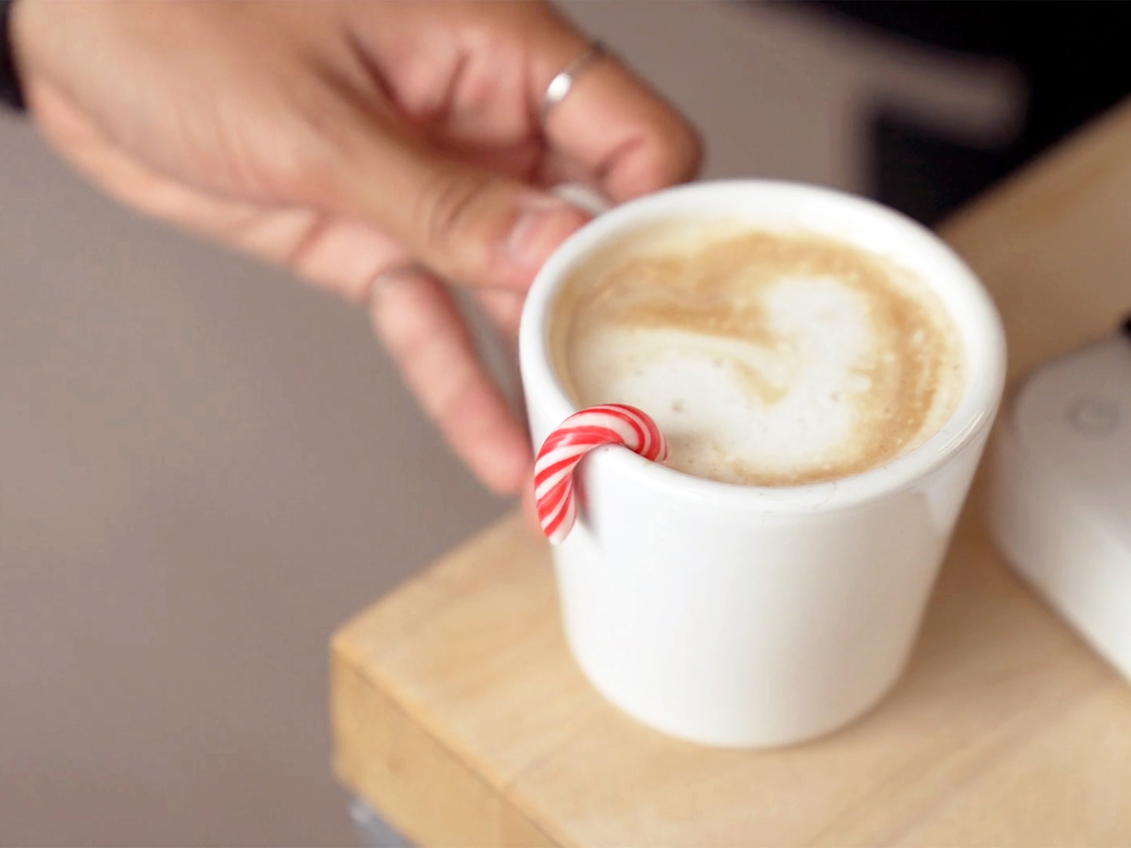 A closeup photo of a hand grabbing a small white mug filled with coffee and foam in the shape of a heart with a candy cane hanging on the rim for garnish.