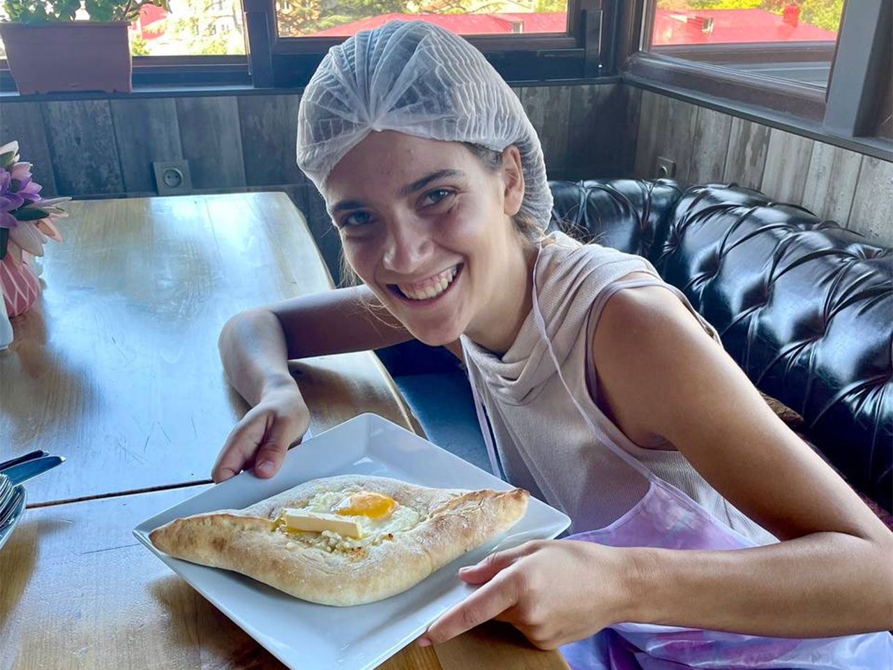 Smiling, UC Davis Russian major Darian Lee sits in a well-lit alcove wearing a hair net. She displays a plate with a boat-shaped pastry filled with cheese.