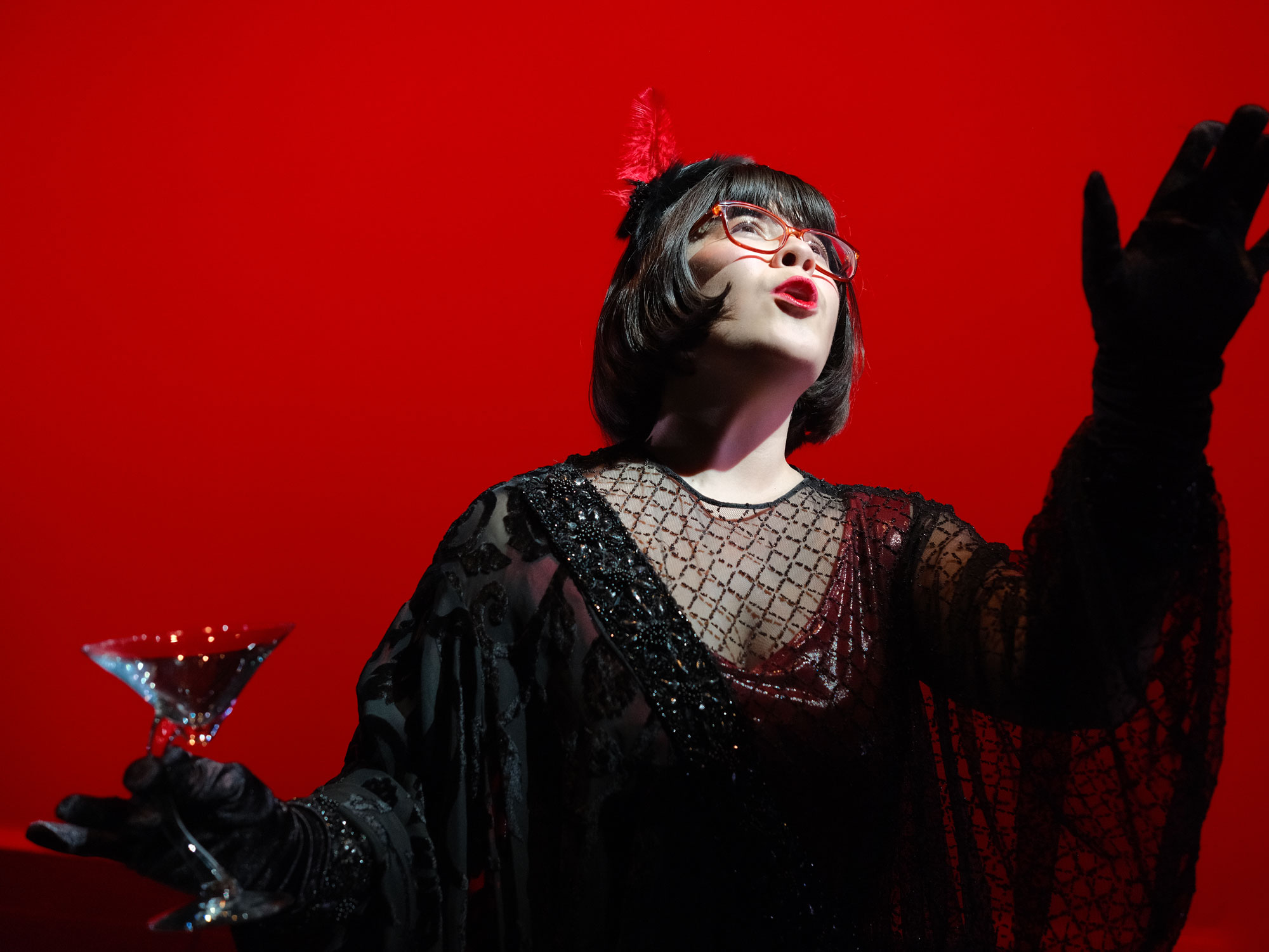 A performer in a black outfit holds a martini glass, set against a vibrant red background.